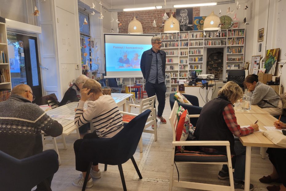 W bibliotece grupa seniorów (8 osób) siedzi przy stołach rozwiązują zadania na kartkach. Dajek po sali chodzi mężczyzna, za nim wyświetlacz z prezentacją.