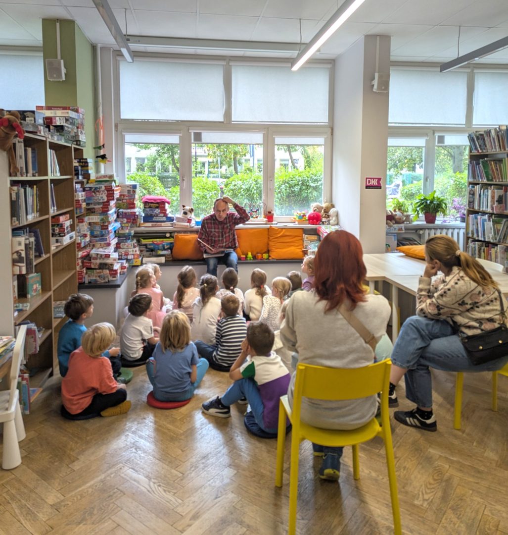 zdjęcie mężczyzny siedzącego i czytającego książkę siedzącym na podłodze dzieciom we wnętrzu biblioteki dla dzieci nr xv. w tle regały biblioteczne.