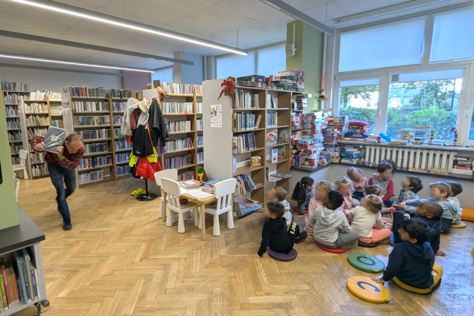 zdjęcie mężczyzny biegnącego z książką do siedzących na podłodze dzieciom we wnętrzu biblioteki dla dzieci nr xv. w tle regały biblioteczne.