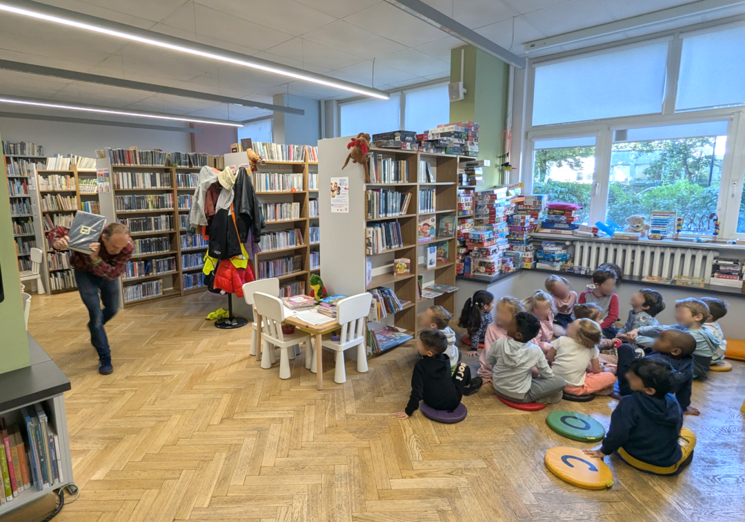 zdjęcie mężczyzny biegnącego z książką do siedzących na podłodze dzieciom we wnętrzu biblioteki dla dzieci nr xv. w tle regały biblioteczne.