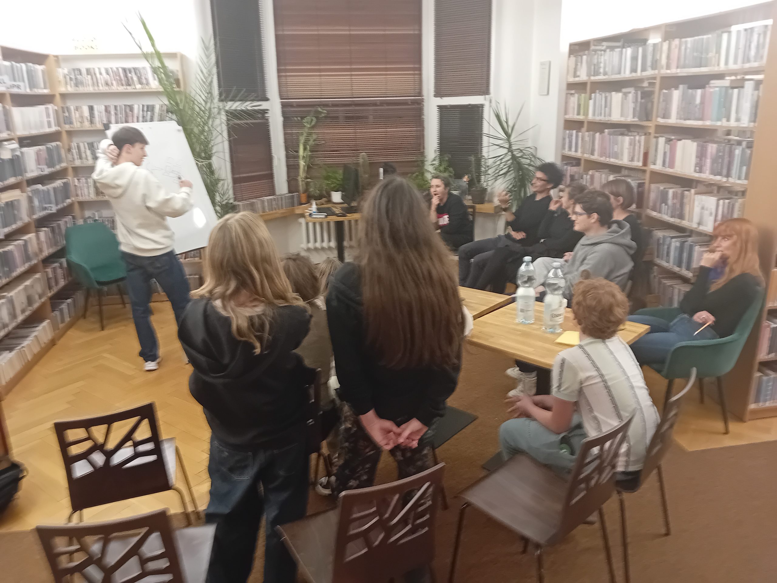 grafika wyróżniająca przedstawiająca uczestników kalamburów literackich. Jeden uczestnik na tablicy rysuje tytuł książki. Pozostali uczestnicy siedzą na krzesełkach wokół niego