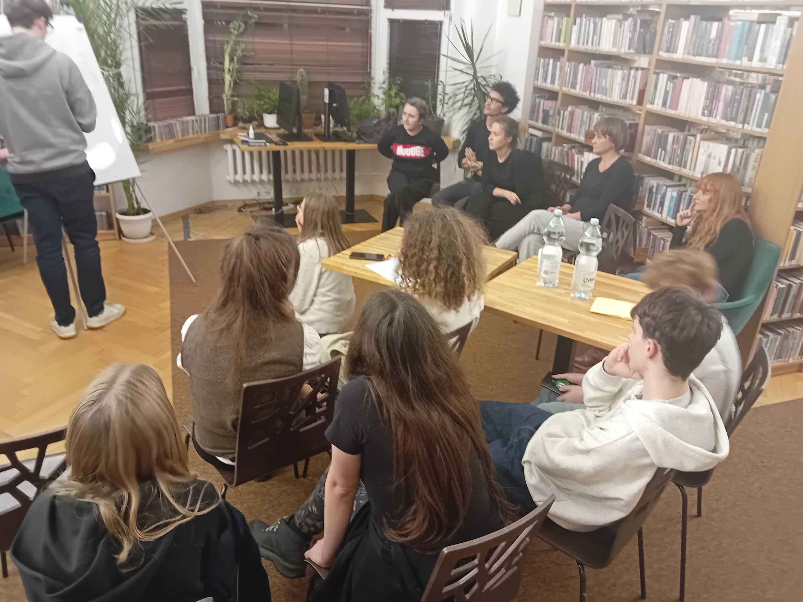 grafika wyróżniająca przedstawiająca uczestników kalamburów literackich. Jeden uczestnik na tablicy rysuje tytuł książki. Pozostali uczestnicy siedzą na krzesełkach wokół niego