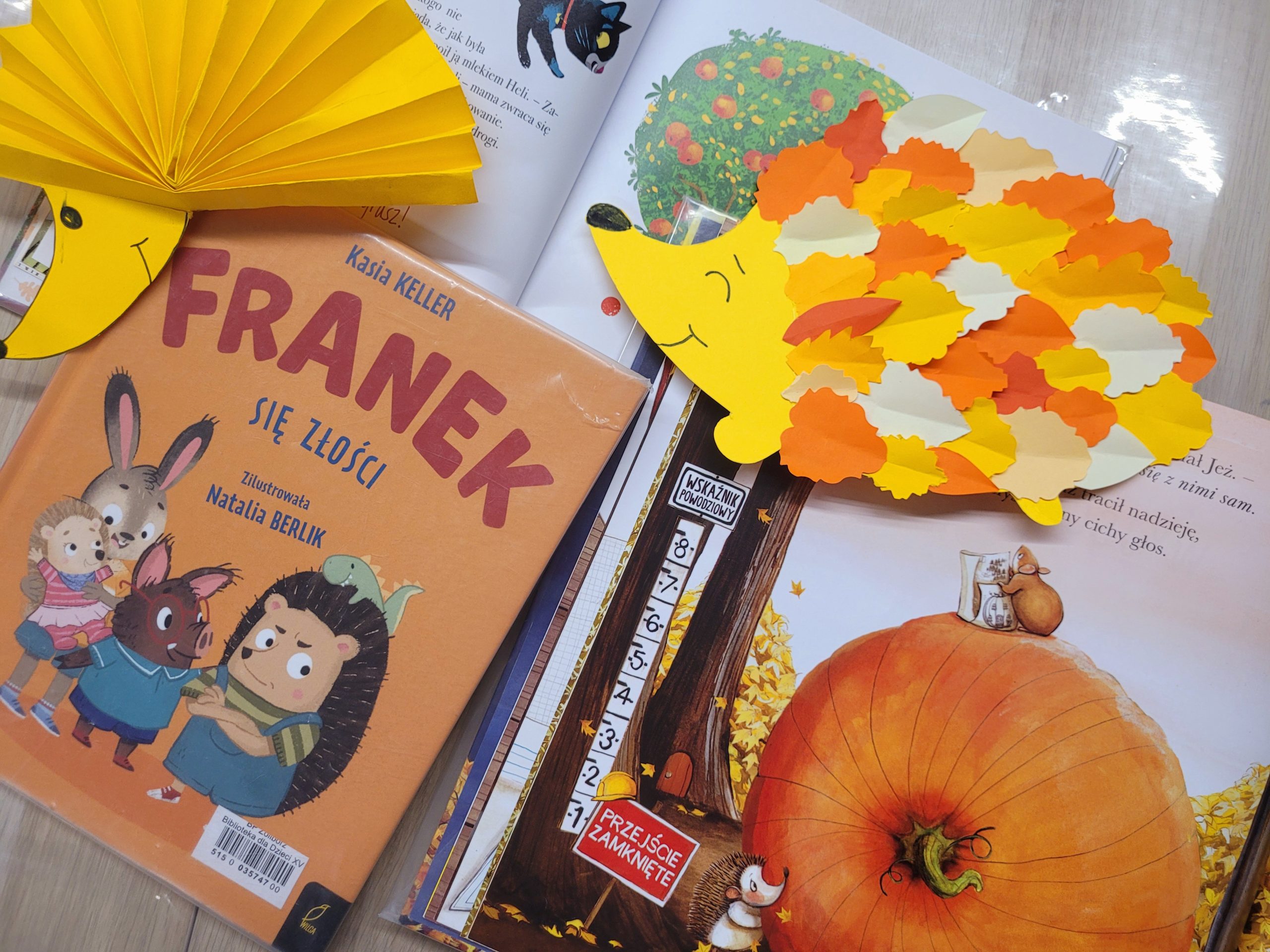 Stolik z książkami i pracami plastycznymi. Na środku książka pt. „Franek się złości” z ilustracją zwierzęcych postaci. Obok otwarte strony innej książki z jesienną ilustracją – wielką dynią, myszką i drzewem. Na książkach leży papierowy jeż wykonany z żółtego papieru i kolorowych, jesiennych listków. W lewym górnym rogu widać wachlarzowo złożony papier imitujący kolce kolejnego jeża.