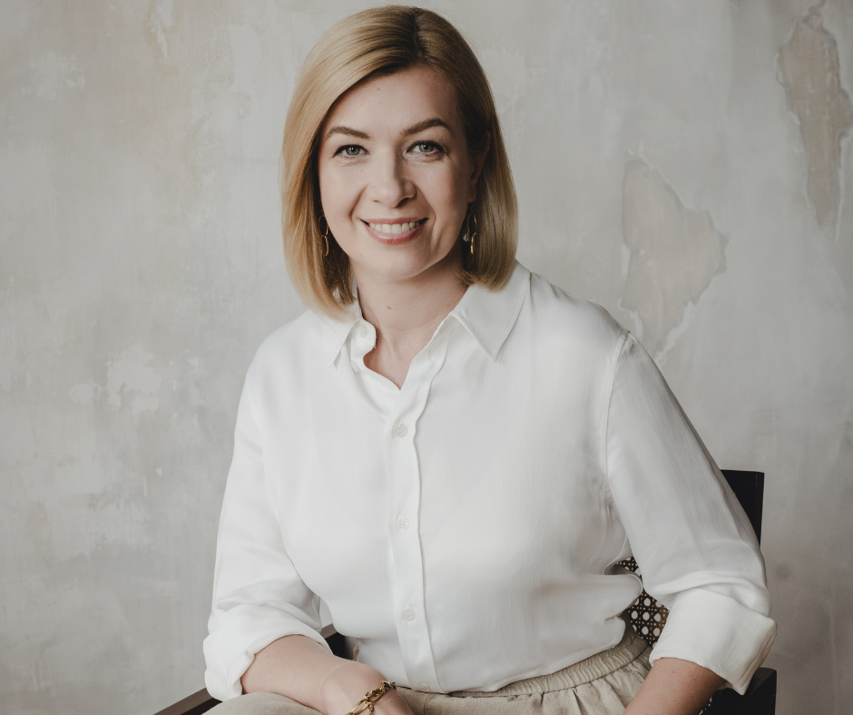 Kobieta siedząca na krześle, uśmiechnięta, ubrana w białą koszulę z podwiniętymi rękawami i jasne spodnie. Ma jasne, proste włosy do ramion. Tło jest jasne, o lekko postarzanej, beżowo-szarej fakturze ściany.