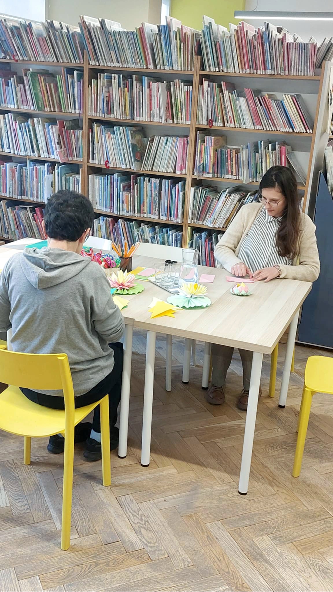 Przy stole dwie osoby wykonują prace plastyczne. Na stole materiały plastyczne, ołówki, kolorowy papier.