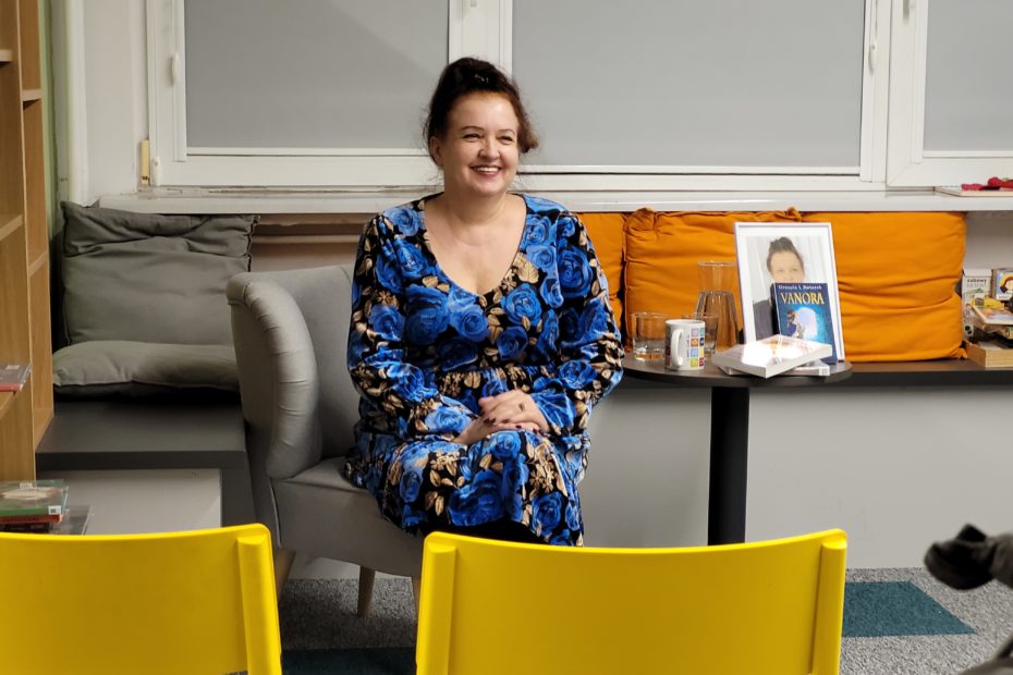 Kobieta w sukience z niebieskim wzorem róż siedzi na szarej kanapie w nowoczesnym wnętrzu biblioteki. Uśmiecha się ciepło, a jej włosy są upięte w luźny kok. W tle widać stolik z książkami, w tym egzemplarz zatytułowany „Vanora", oraz szklankę z wodą. Przed nią stoją żółte plastikowe krzesła.