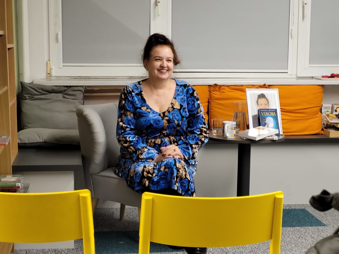 Kobieta w sukience z niebieskim wzorem róż siedzi na szarej kanapie w nowoczesnym wnętrzu biblioteki. Uśmiecha się ciepło, a jej włosy są upięte w luźny kok. W tle widać stolik z książkami, w tym egzemplarz zatytułowany „Vanora", oraz szklankę z wodą. Przed nią stoją żółte plastikowe krzesła.