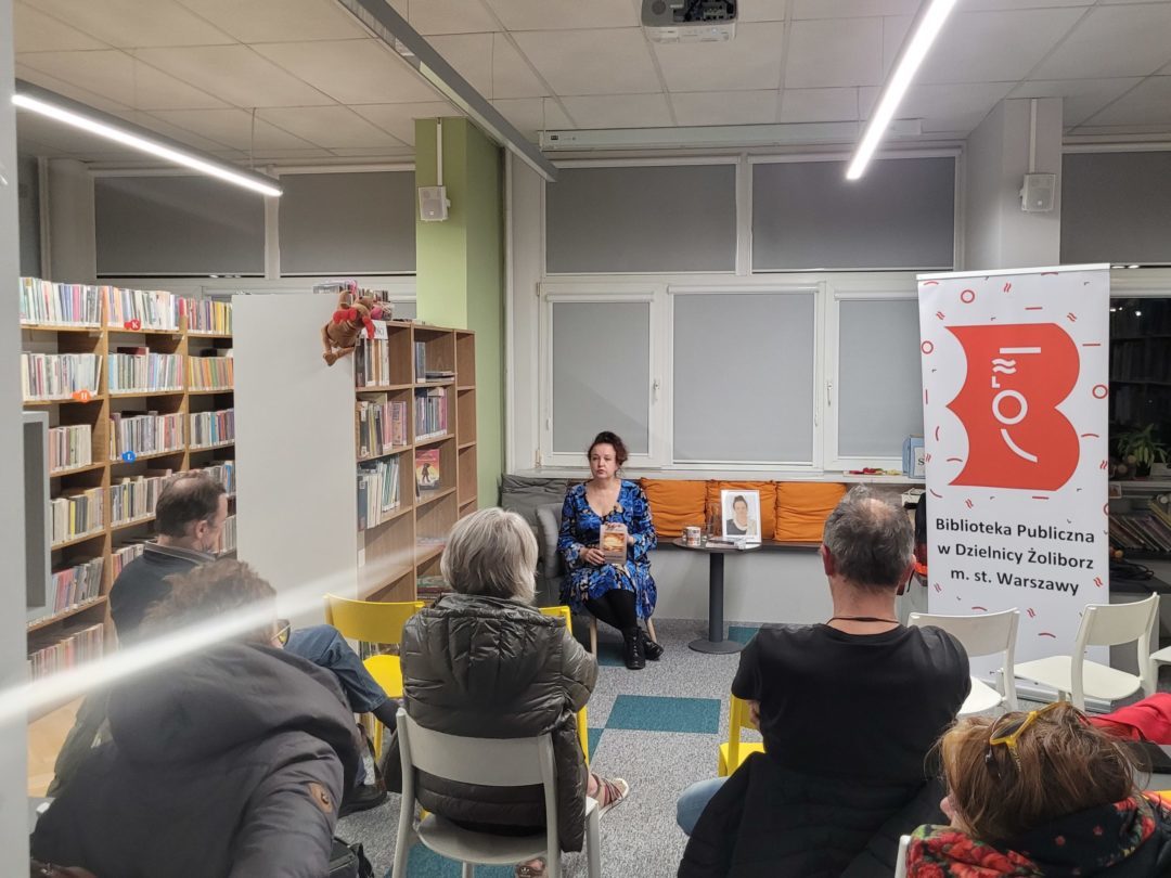 Spotkanie autorskie w Bibliotece Publicznej w Dzielnicy Żoliborz m. st. Warszawy. Autorka w niebieskiej sukience siedzi na pomarańczowej kanapie, prezentując swoją książkę przed grupą słuchaczy. Uczestnicy wydarzenia siedzą na kolorowych krzesłach w przestrzeni bibliotecznej wypełnionej regałami z książkami. W tle widoczny jest baner biblioteki