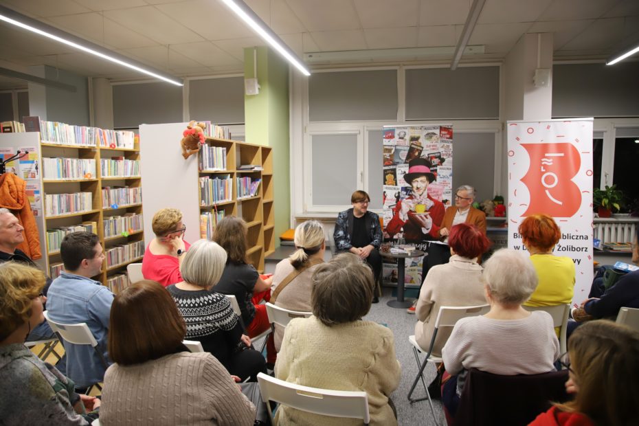 Spotkanie autorskie w bibliotece widziane od tyłu, z perspektywy publiczności. W pierwszym rzędzie siedzi grupa osób zwrócona w stronę prowadzących. Przed nimi, na tle planszy z ilustracją i baneru biblioteki, siedzą dwie osoby prowadzące rozmowę. Otoczenie to jasna czytelnia z regałami wypełnionymi książkami.