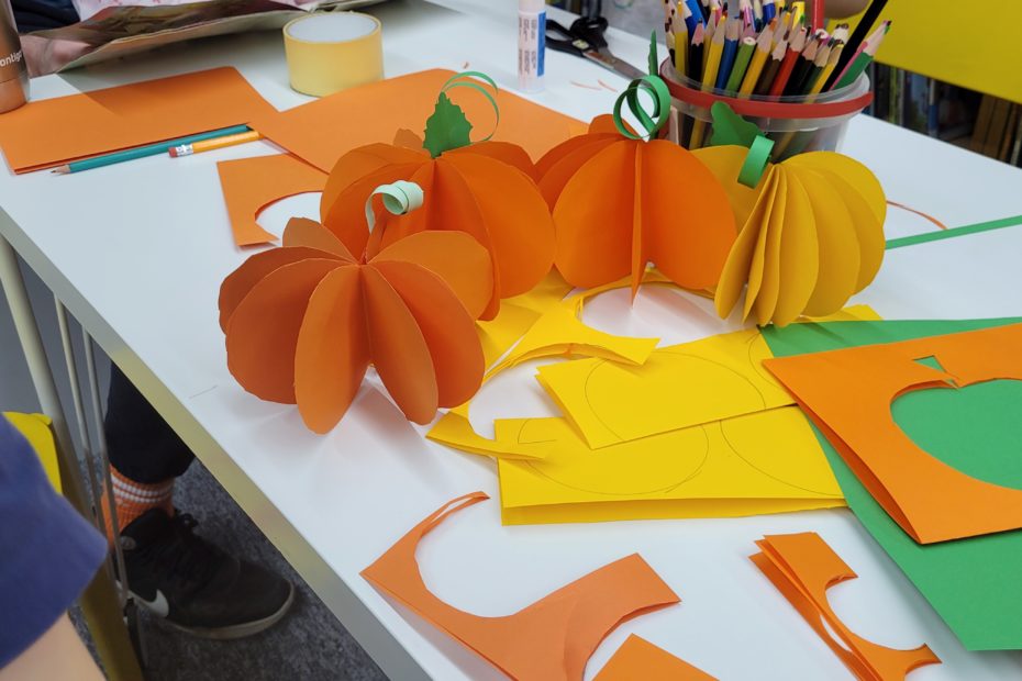 Grupa dzieci i dorosłych wykonuje prace plastyczne w bibliotece - widać tylko ręce. Dzieci siedzą przy stołach, wycinając i klejąc kolorowe kartki, a dorośli pomagają im w zajęciach. Na stole pomarańczowe i żółte dynie z papieru, kartki, kleje, kredki.