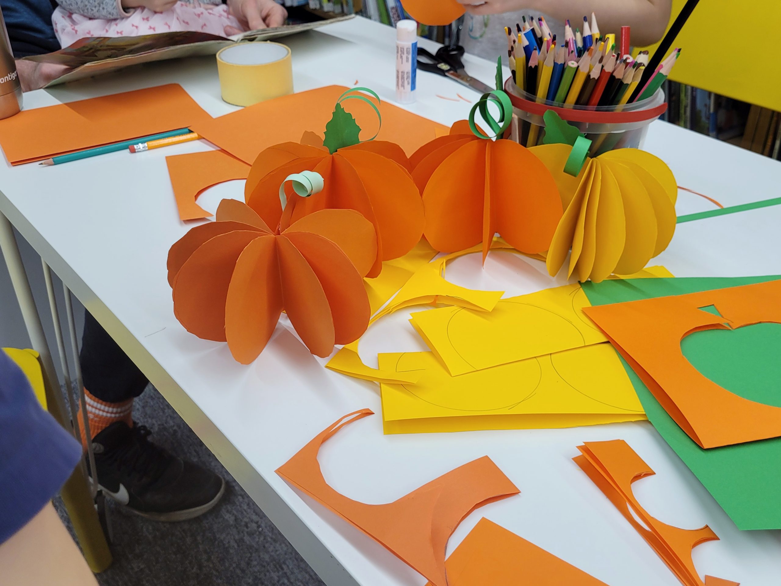 Grupa dzieci i dorosłych wykonuje prace plastyczne w bibliotece - widać tylko ręce. Dzieci siedzą przy stołach, wycinając i klejąc kolorowe kartki, a dorośli pomagają im w zajęciach. Na stole pomarańczowe i żółte dynie z papieru, kartki, kleje, kredki.