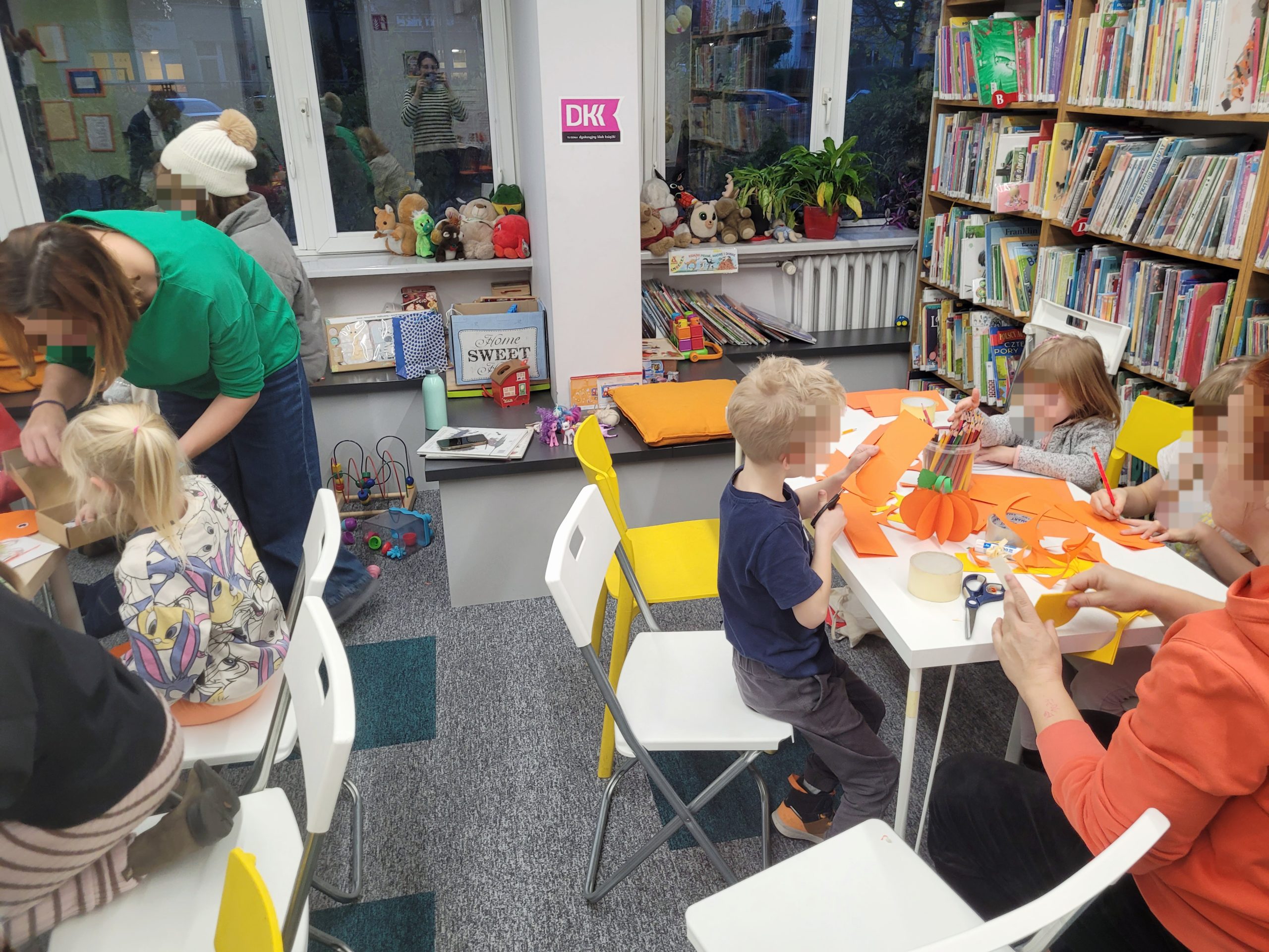 Grupa dzieci i dorosłych wykonuje prace plastyczne w bibliotece. Dzieci siedzą przy stołach, wycinając i klejąc kolorowe kartki, a dorośli pomagają im w zajęciach. W tle widać półki z książkami, pluszowe zabawki oraz duże okna.