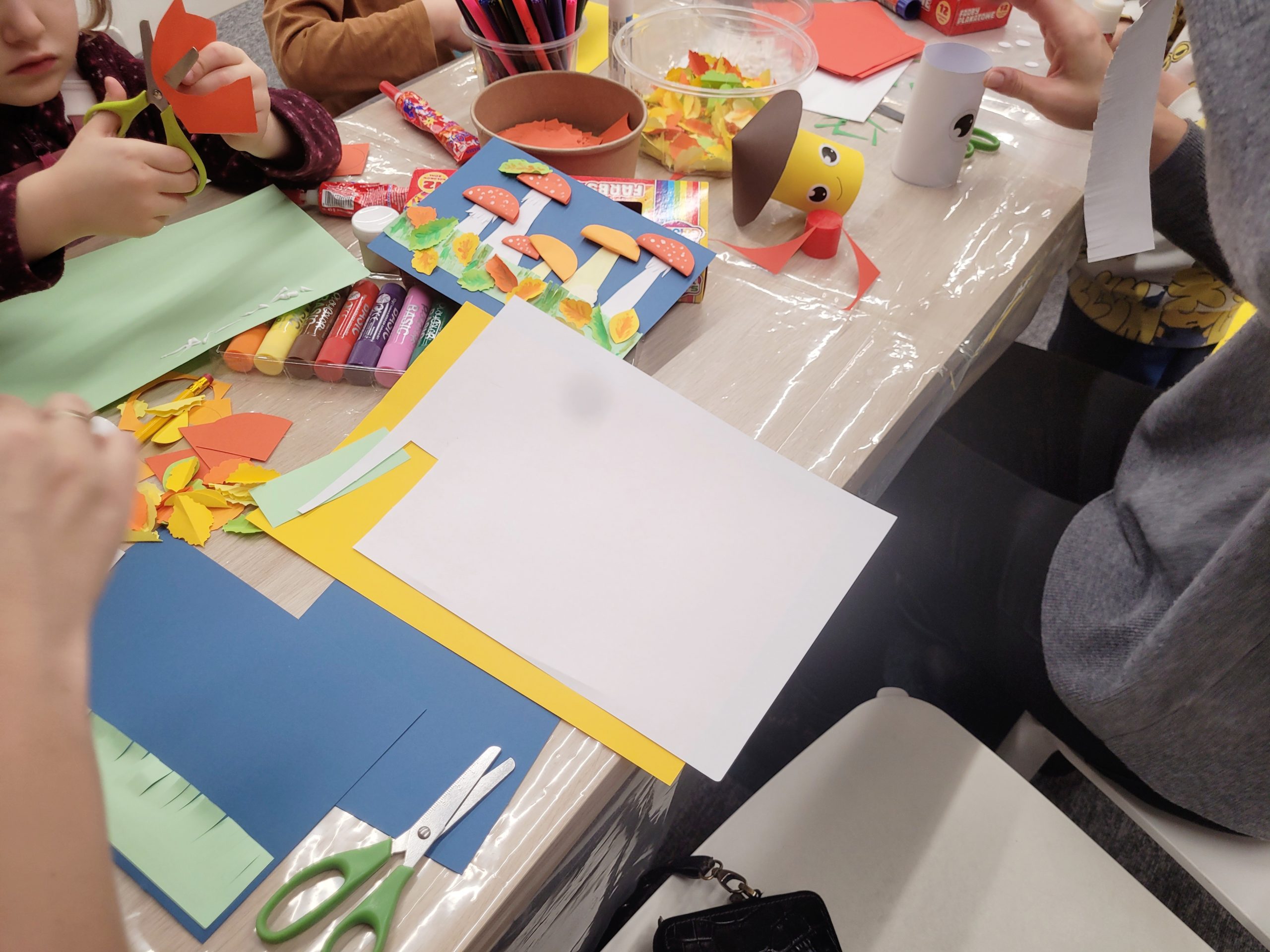 Dzieci siedzą przy stole pełnym materiałów plastycznych, takich jak kolorowy papier, klej, nożyczki i kredki. Jedno z dzieci wycina elementy z pomarańczowego papieru, a na stole leżą przygotowane dekoracje w kształcie grzybków oraz jesienne liście.