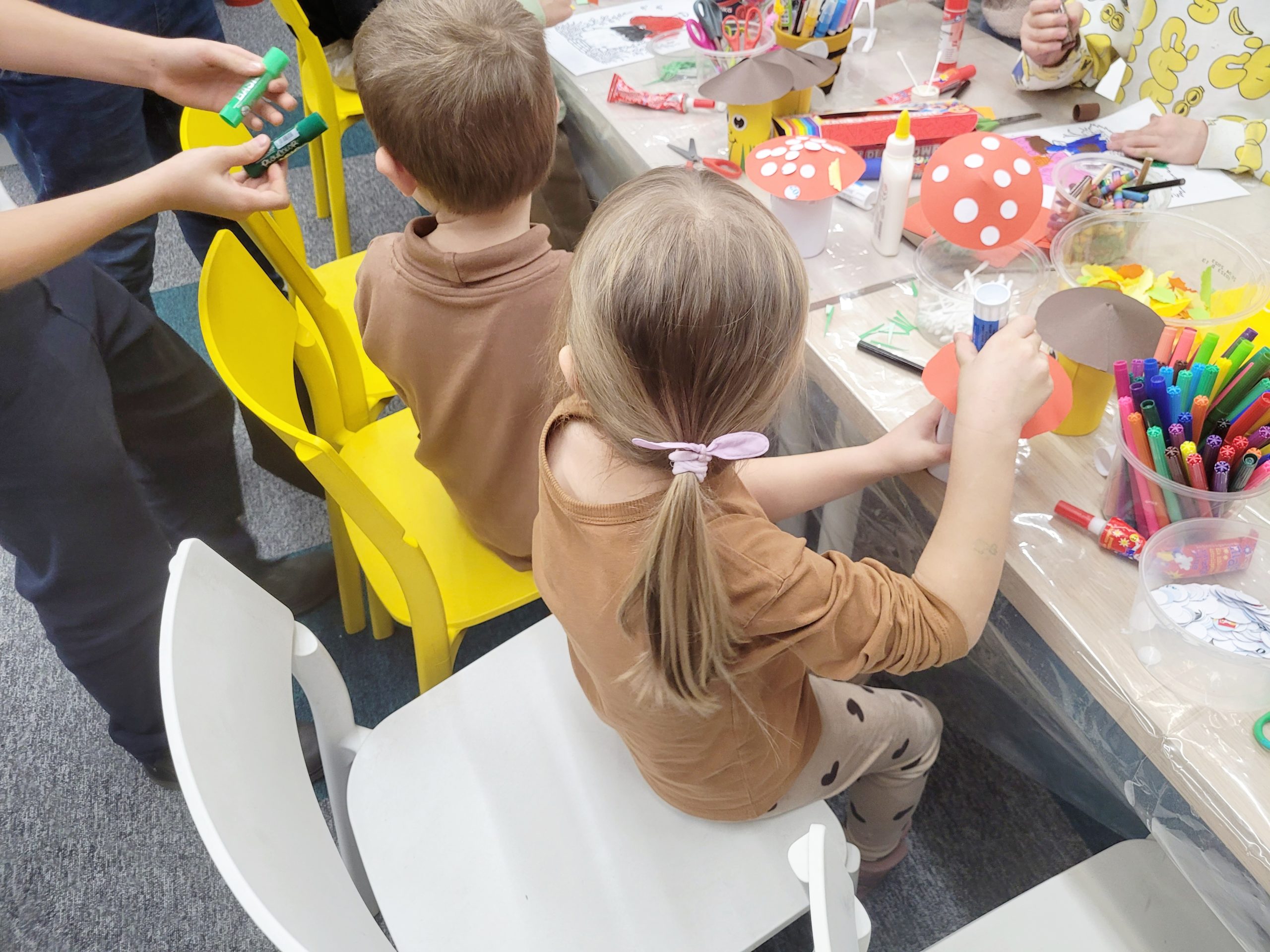 Dzieci siedzą przy stole pełnym materiałów plastycznych, takich jak kolorowy papier, klej, nożyczki i kredki. Dzieci wykonują kolorowe grzyby.