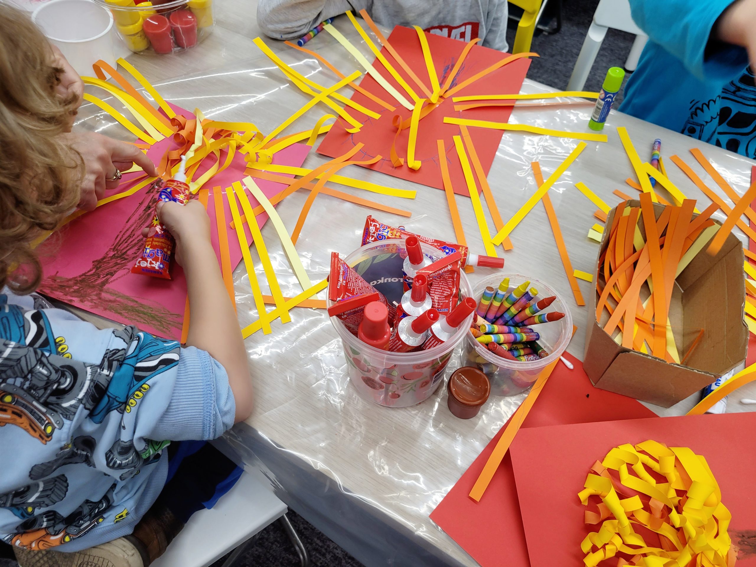 Dzieci tworzą kolorowe prace plastyczne z papierowych pasków w odcieniach żółci, pomarańczy i czerwieni, które przypominają promienie słońca lub płatki kwiatów. Na stole widoczne są materiały do rękodzieła, w tym kleje w pojemnikach, kredki oraz kolorowe papiery rozłożone na folii ochronnej.