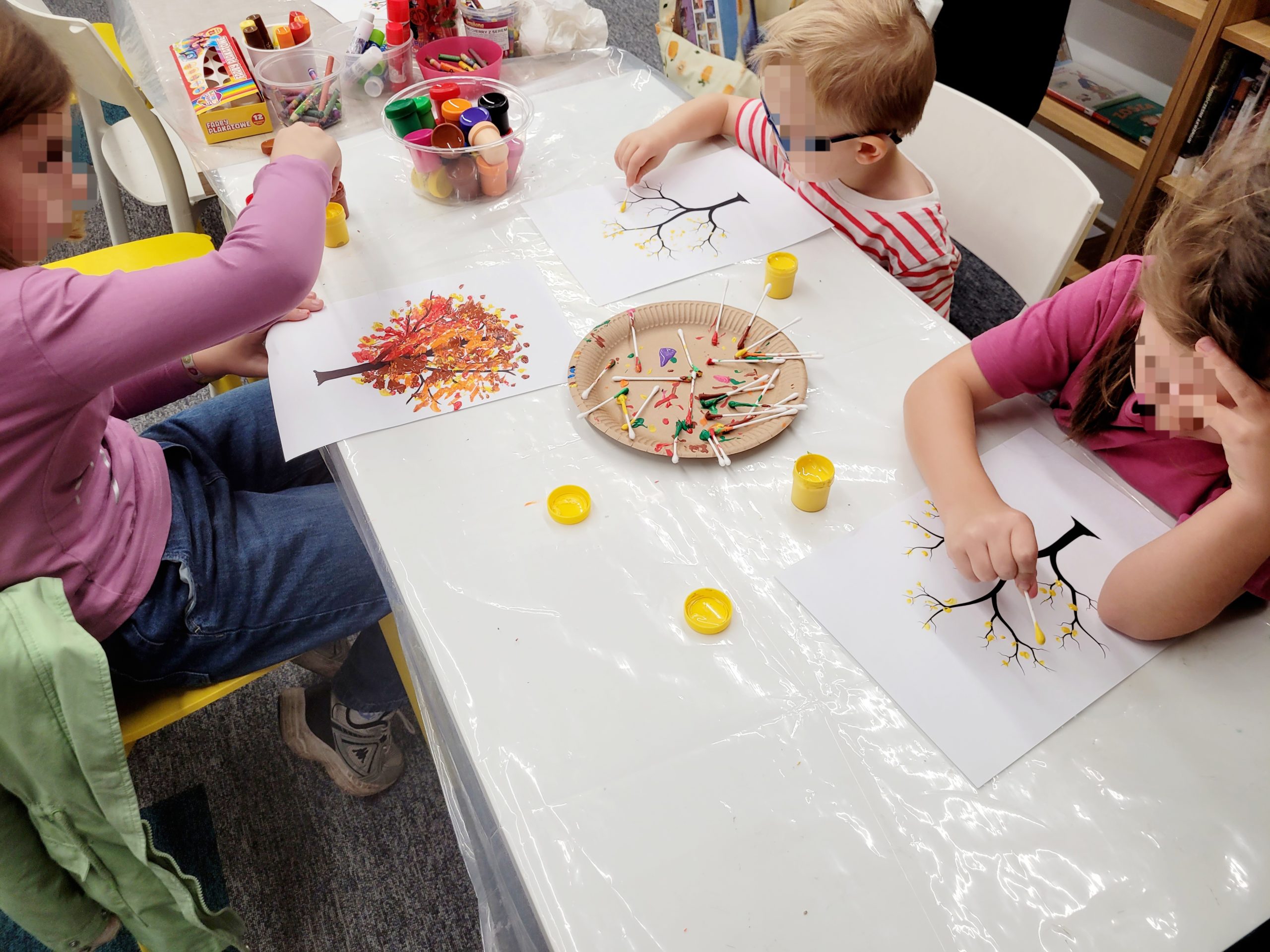 Dzieci tworzą jesienne prace plastyczne, malując drzewa .Na stole widoczne są farby, kleje w sztyfcie oraz różnokolorowe materiały do rękodzieła rozłożone na folii ochronnej.