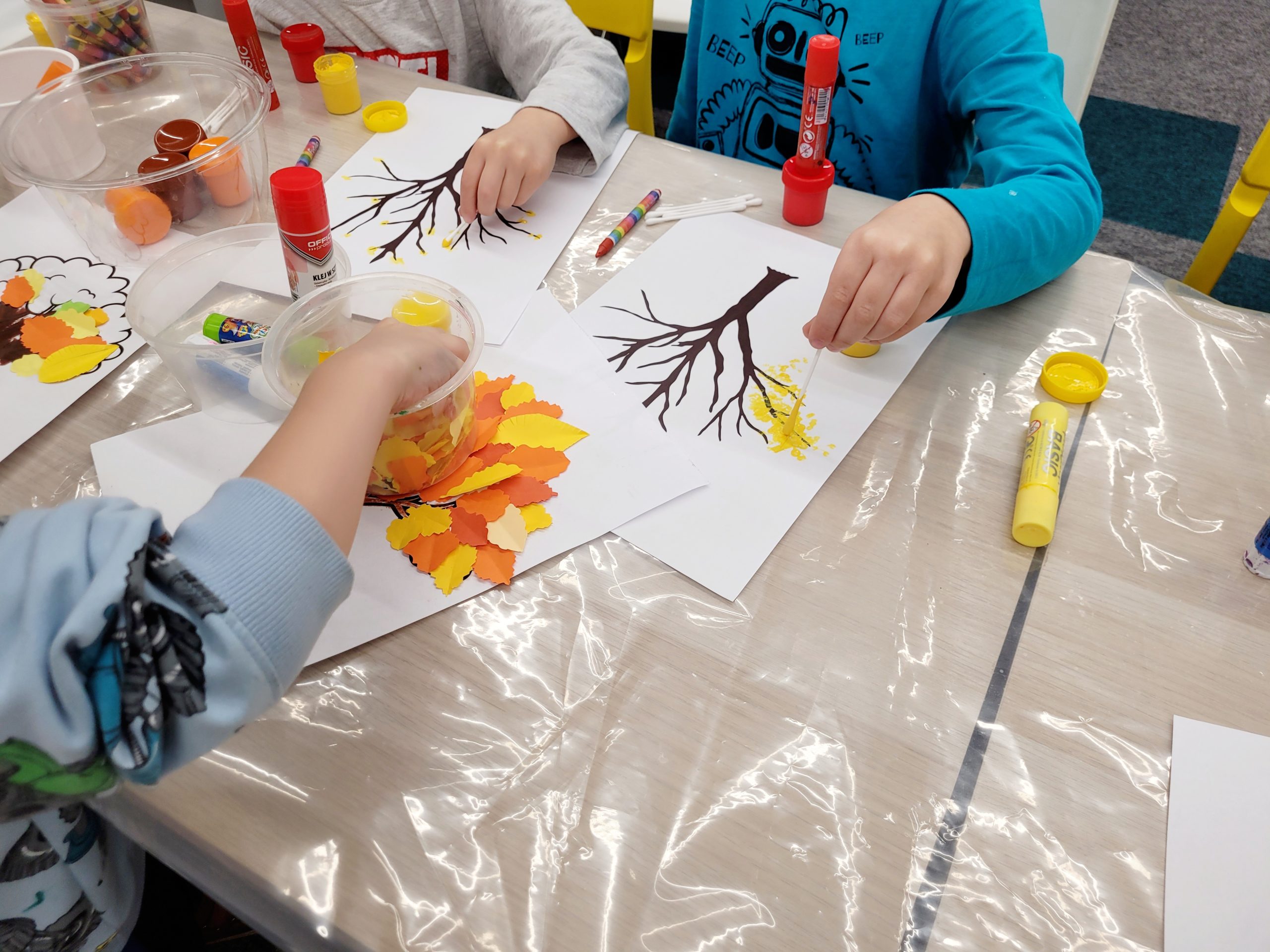 Dzieci tworzą jesienne prace plastyczne, malując drzewa i przyklejając kolorowe liście z papieru w odcieniach żółci, pomarańczy i czerwieni. Na stole widoczne są farby, kleje w sztyfcie oraz różnokolorowe materiały do rękodzieła rozłożone na folii ochronnej.