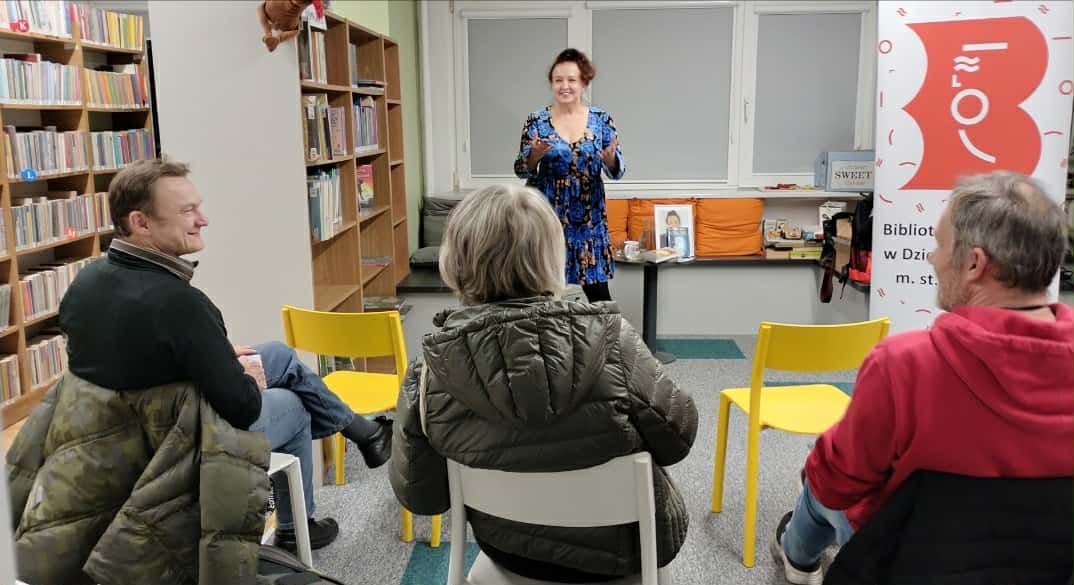 Spotkanie autorskie w Bibliotece Publicznej w Dzielnicy Żoliborz m. st. Warszawy. Autorka w niebieskiej sukience stoi przed grupą słuchaczy. Uczestnicy wydarzenia siedzą na kolorowych krzesłach w przestrzeni bibliotecznej wypełnionej regałami z książkami. W tle widoczny jest baner biblioteki.