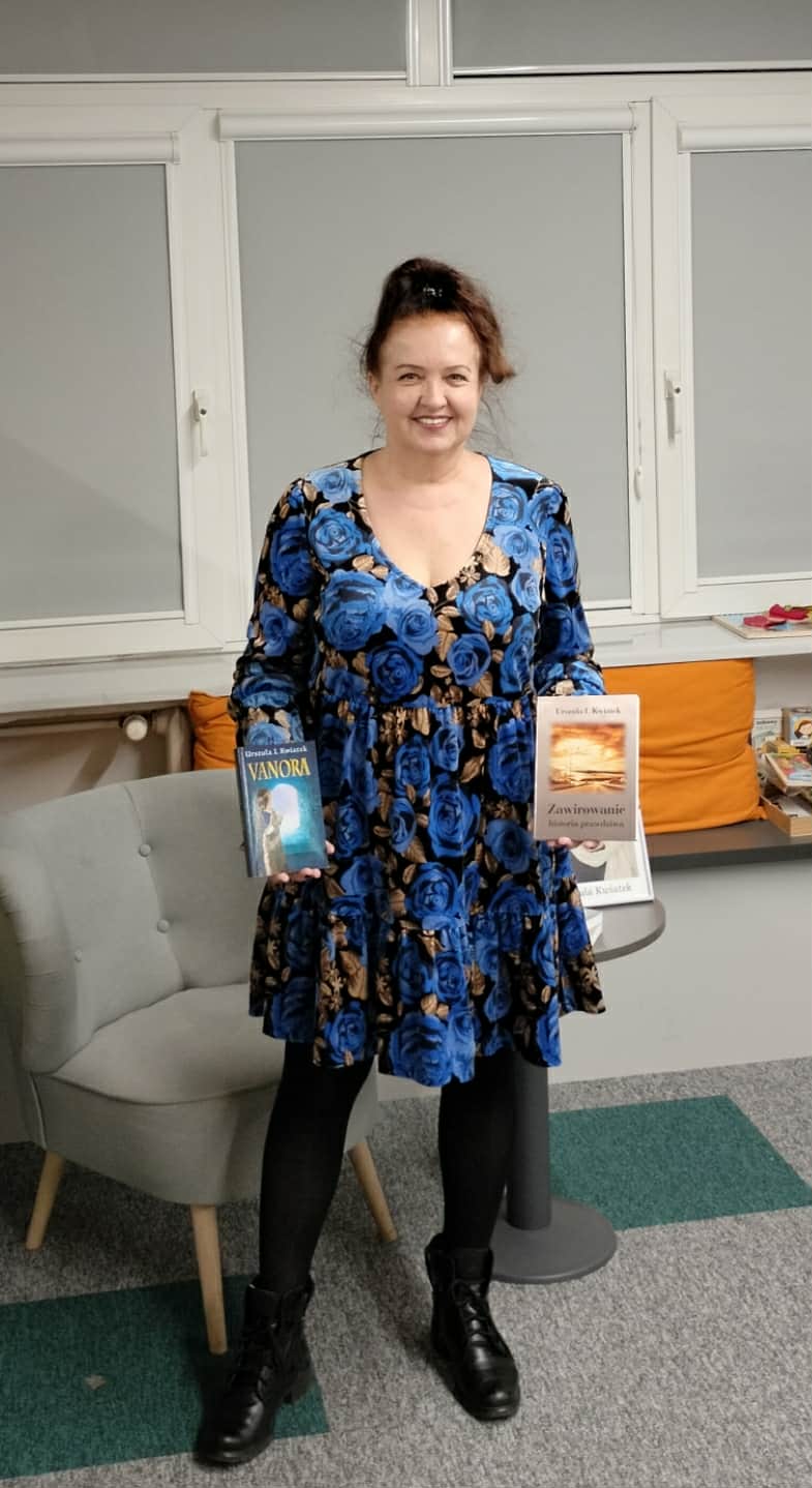 Uśmiechnięta kobieta w sukience z niebieskim wzorem róż stoi w bibliotece lub księgarni, trzymając w rękach dwie książki. Na zdjęciu widać jedną z publikacji zatytułowaną „Vanora" oraz drugą książkę z napisem „Zawirowanie".