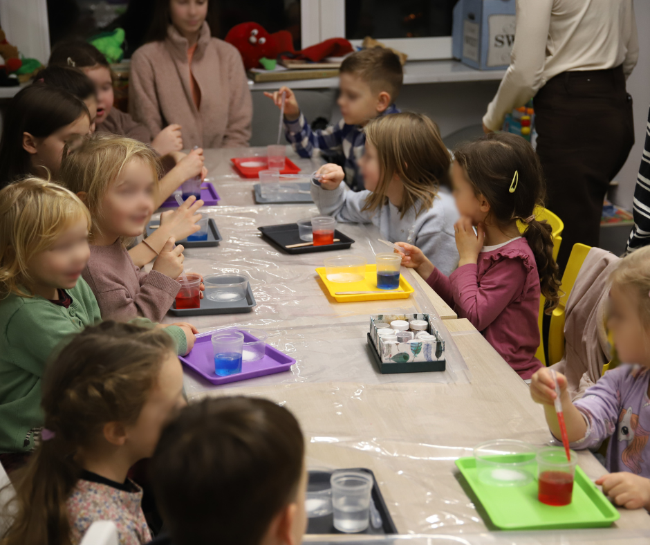 Grupa dzieci przy stołach robią eksperymenty, mają tacki przed sobą w różnych kolorach (żółte, szare, fioletowe, czerwone, zielone), na których są przezroczyste naczynka z płynem.