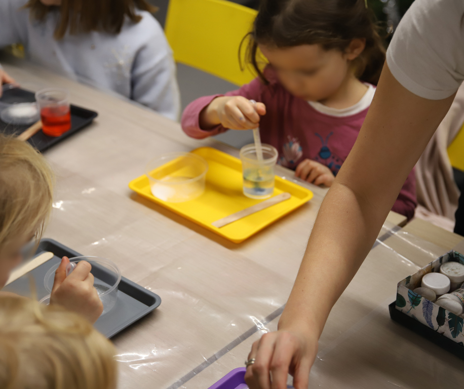 Grupa dzieci przy stołach, mają tacki przed sobą w różnych kolorach (żółta, szara, czarna), na których są przezroczyste naczynka. Dzieci mieszają płyny.