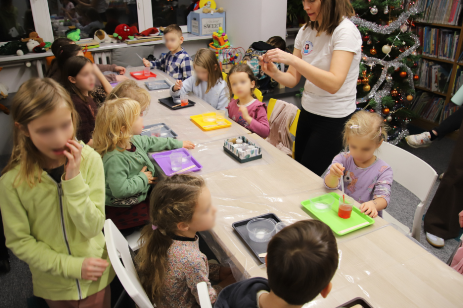 Grupa dzieci przy stołach robią eksperymenty, mają tacki przed sobą w różnych kolorach (żółte, szare, fioletowe, czerwone, zielone), na których są przezroczyste naczynka z płynem. Obok stołu stoi kobieta w białej koszulce i coś pokazuje dzieciom. W tle choinka.