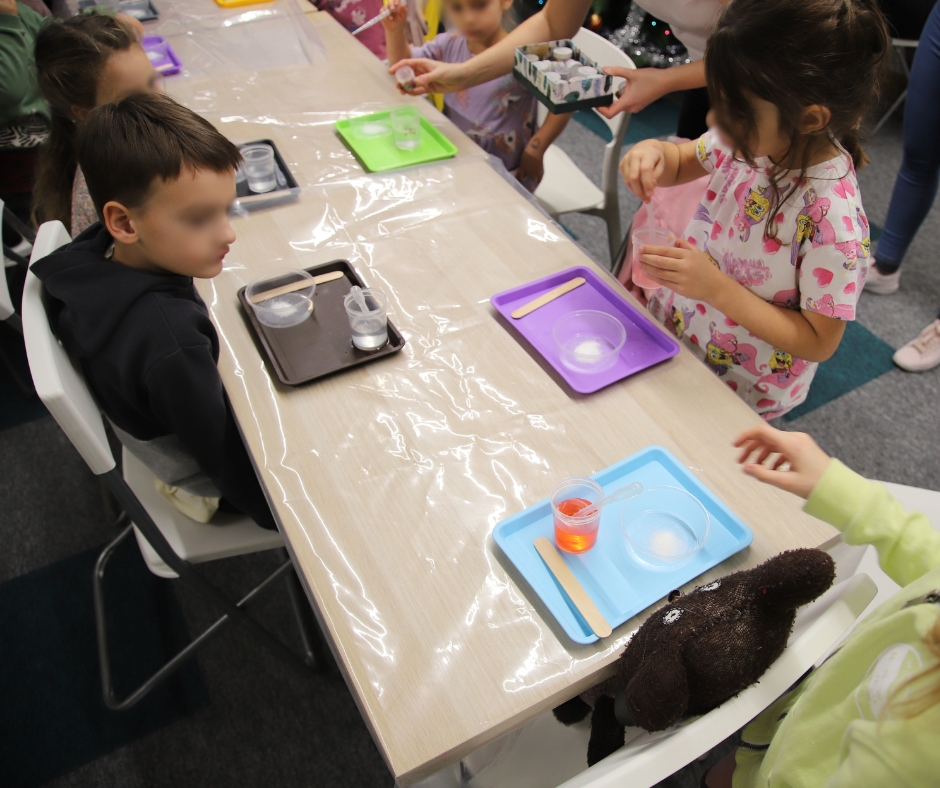 Grupa dzieci przy stołach robią eksperymenty, mają tacki przed sobą w różnych kolorach (zielone, fioletowe, czarne, niebieskie), na których są przezroczyste naczynka z płynem.