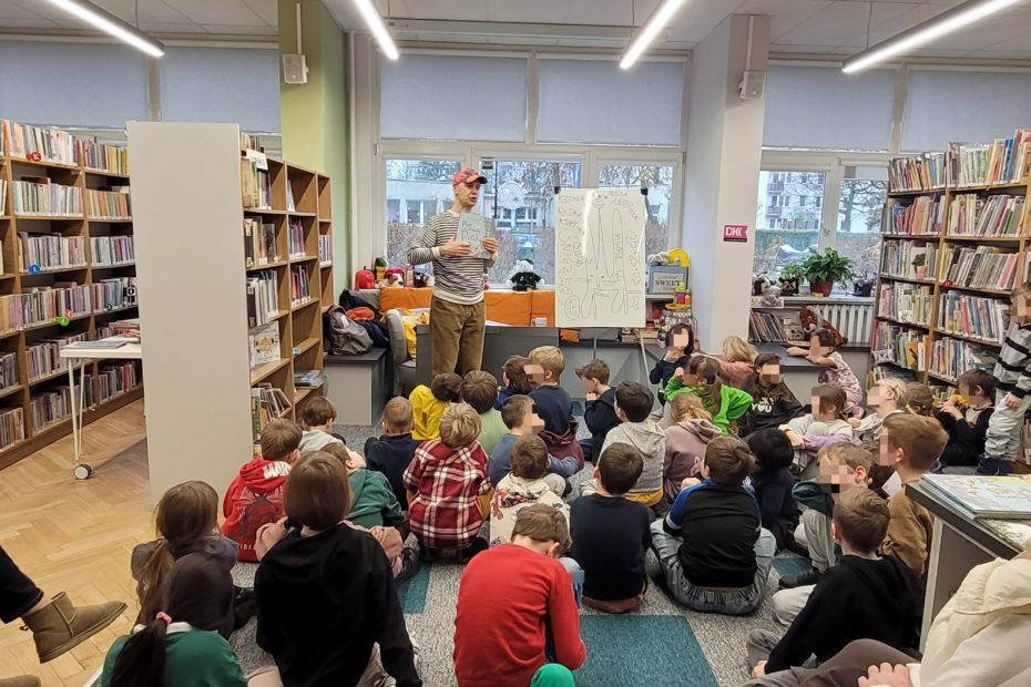 Grupa dzieci siedzi na podłodze w czytelni biblioteki i słucha prowadzącego w czapce i paskowanej bluzie, który stoi przy sztaludze na której są rysunki markerem. Prowadzący trzyma w rękach książkę z jasną okładką. Wokół widać regały pełne książek i jasne, duże okna.