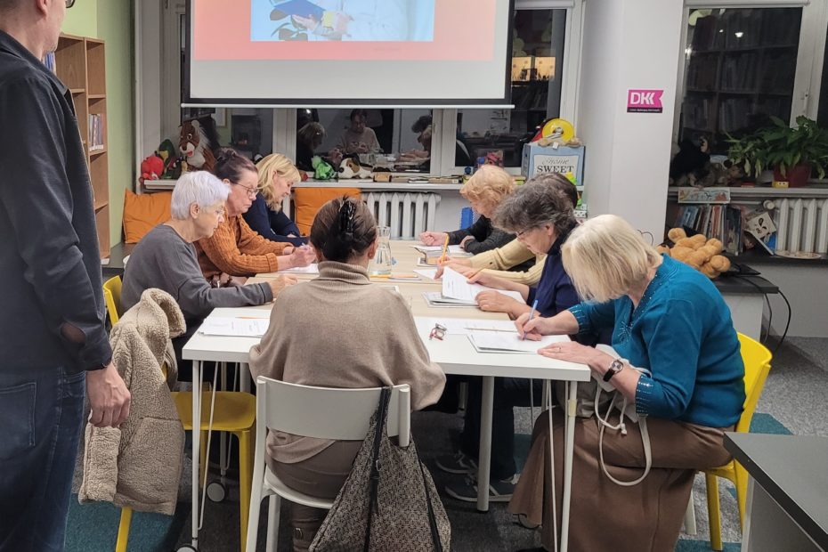 Grupa starszych osób siedzi przy stoliku w bibliotece i wykonuje ćwiczenia pisemne podczas zajęć dotyczących pamięci i koncentracji. Na ekranie projektora widoczny jest slajd z tytułem ‘Pamięć i koncentracja’. Prowadzący stoi obok stołu