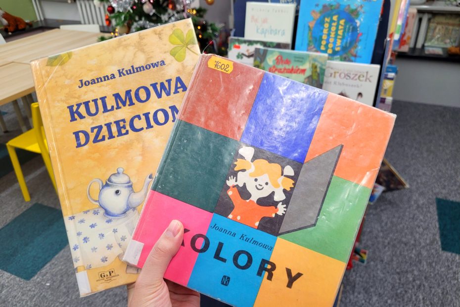 Dwie książki Joanny Kulmowej trzymane w dłoni: „Kulmowa dzieciom” oraz „Kolory”, na tle biblioteki dziecięcej z regałami książek i świąteczną choinką w tle.