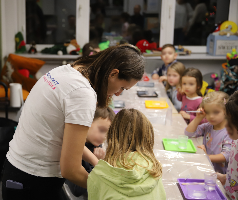 Grupa dzieci przy stołach robią eksperymenty, mają tacki przed sobą w różnych kolorach (żółte, szare, fioletowe, czerwone, zielone), na których są przezroczyste naczynka z płynem. Na pierwszym planie schylona kobieta w białej koszulce.