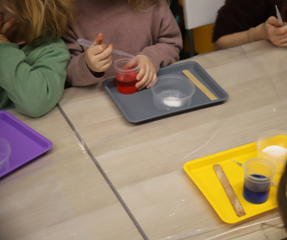 Grupa dzieci przy stołach robią eksperymenty, mają tacki przed sobą w różnych kolorach (żółta, szara, fioletowa), na których są przezroczyste naczynka z płynem.