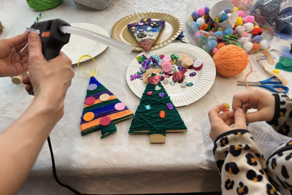 grafika przedstawiająca ręce dorosłego i dziecka w trakcie zajęć z robienia tekturowych choinek, które ozdabiano kolorową włóczką