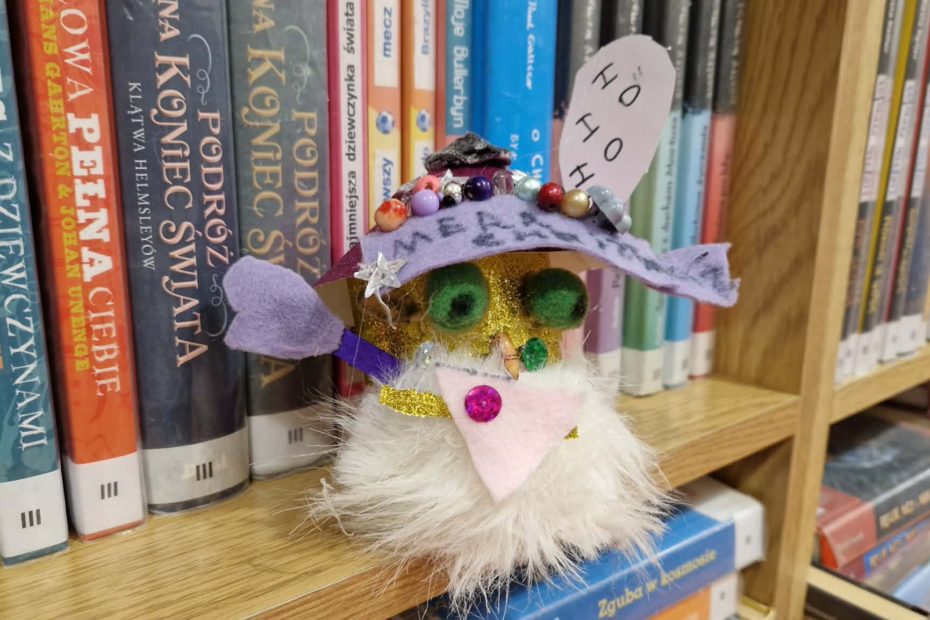 ilustracja przedstawiająca kolorowego skrzata wykonanego ze skarpetki. Ma białą brodę, zielone oczy i fioletową czapkę z filcu przyozdobioną koralikami. Nad czapką jest karteczka z białego papieru z napisem "ho ho"