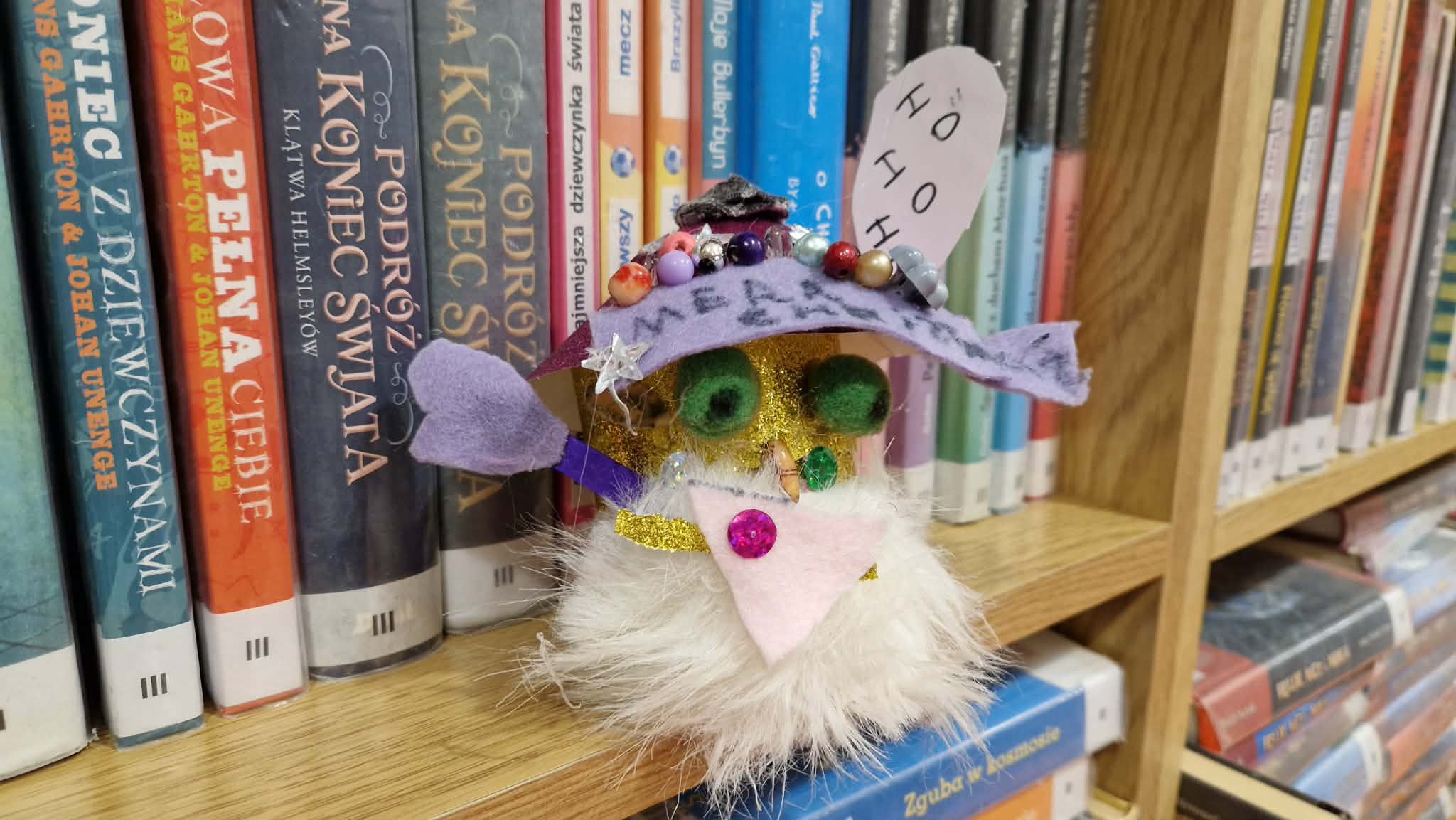 ilustracja przedstawiająca kolorowego skrzata wykonanego ze skarpetki. Ma białą brodę, zielone oczy i fioletową czapkę z filcu przyozdobioną koralikami. Nad czapką jest karteczka z białego papieru z napisem "ho ho"