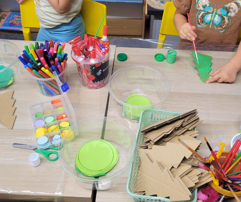 Widok z góry na stół warsztatowy pełen materiałów plastycznych: farby, pędzle, flamastry, nożyczki i papierowe choinki z kartonu. Dzieci malują zielone elementy farbą, dekorując je według własnych pomysłów. Przestrzeń jest uporządkowana i przygotowana do pracy twórczej.