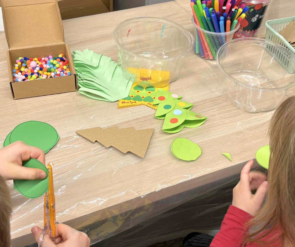 Zbliżenie na stół podczas zajęć plastycznych. Dzieci wycinają zielone papierowe elementy choinek, używając nożyczek. Na stole leżą kolorowe pompony w pudełku, papierowe kształty, flamastry w kubkach oraz plastikowe pojemniki na materiały. Widoczne są dziecięce dłonie pracujące nad dekoracjami.