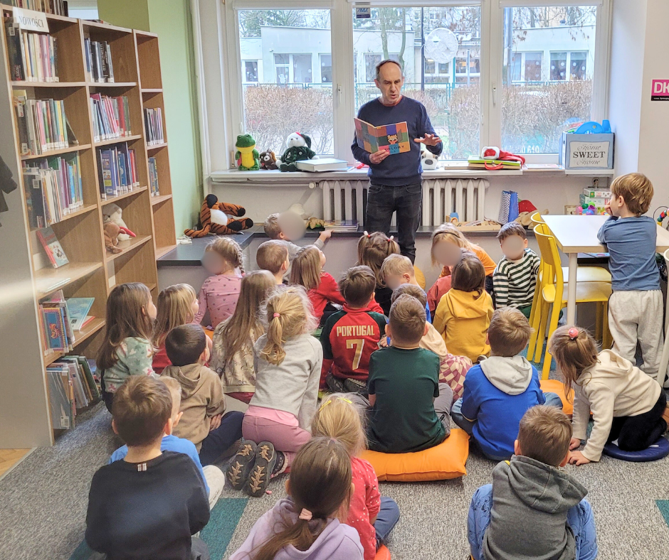Bibliotekarz czyta książkę grupie przedszkolaków siedzących na dywanie w sali bibliotecznej. Dzieci słuchają w skupieniu, w tle widoczne są okna, stoliki, krzesła oraz zabawki, tworzące przyjazną przestrzeń do wspólnego czytania.