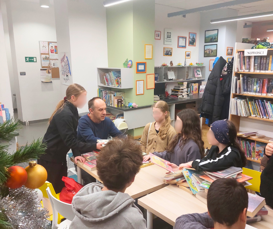 Grupa dzieci i dorosły prowadzący siedzą przy stole w bibliotece, rozmawiają o książkach i pokazują sobie nawzajem wybrane tytuły podczas spotkania Młodzieżowego Dyskusyjnego Klubu Książki.