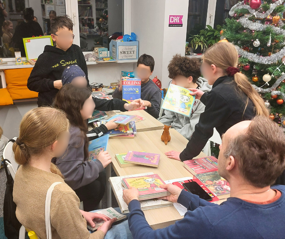 Uczestnicy Młodzieżowego Dyskusyjnego Klubu Książki stoją i siedzą wokół stołu pełnego książek w bibliotece; dzieci prezentują okładki i dyskutują, w tle widoczna choinka świąteczna.