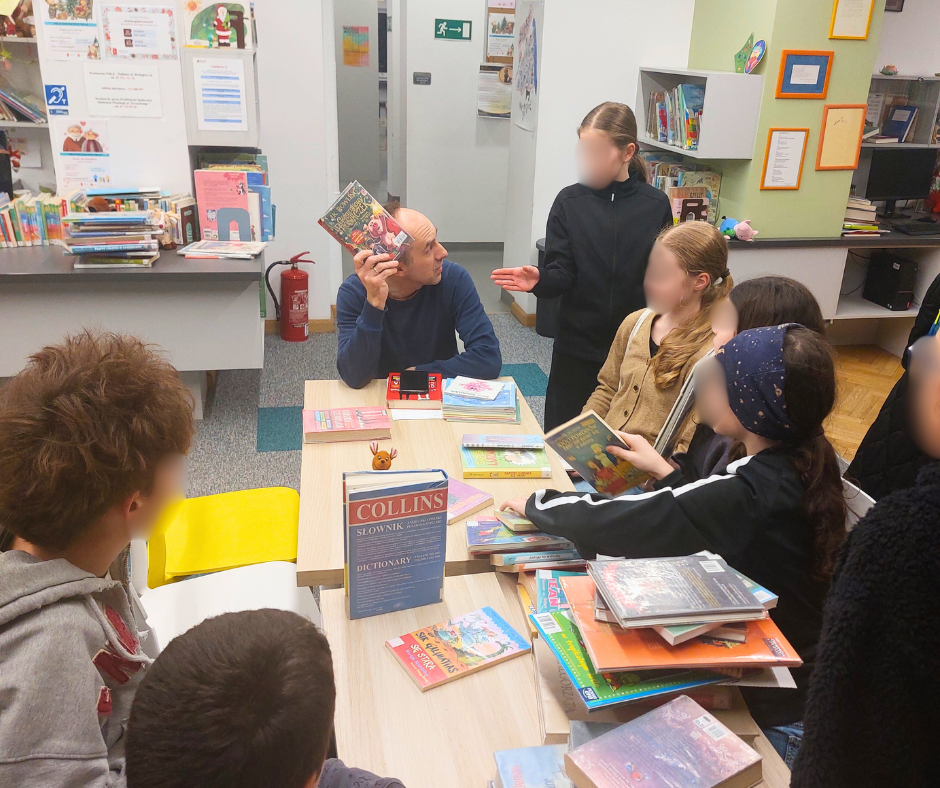 Grupa dzieci i dorosły prowadzący siedzą przy stole w bibliotece, rozmawiają o książkach i pokazują sobie nawzajem wybrane tytuły podczas spotkania Młodzieżowego Dyskusyjnego Klubu Książki.