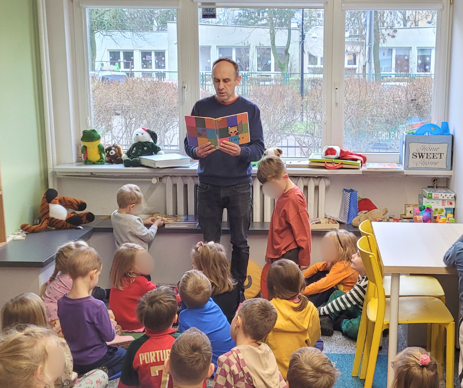 Bibliotekarz czyta książkę grupie przedszkolaków siedzących na dywanie w sali bibliotecznej. Dzieci słuchają w skupieniu, w tle widoczne są okna, stoliki, krzesła oraz zabawki, tworzące przyjazną przestrzeń do wspólnego czytania.