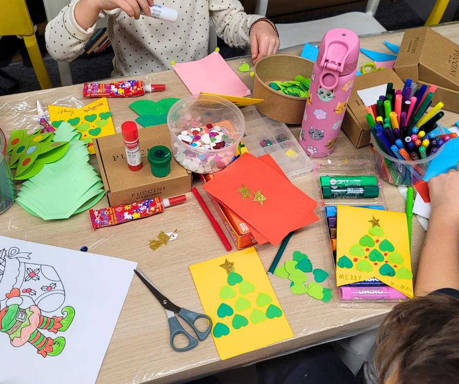 Widok z góry na stół warsztatowy pełen materiałów plastycznych: farby, pędzle, flamastry, nożyczki i papierowe choinki z kartonu. Dzieci kleją choinki z zielonych serduszek.