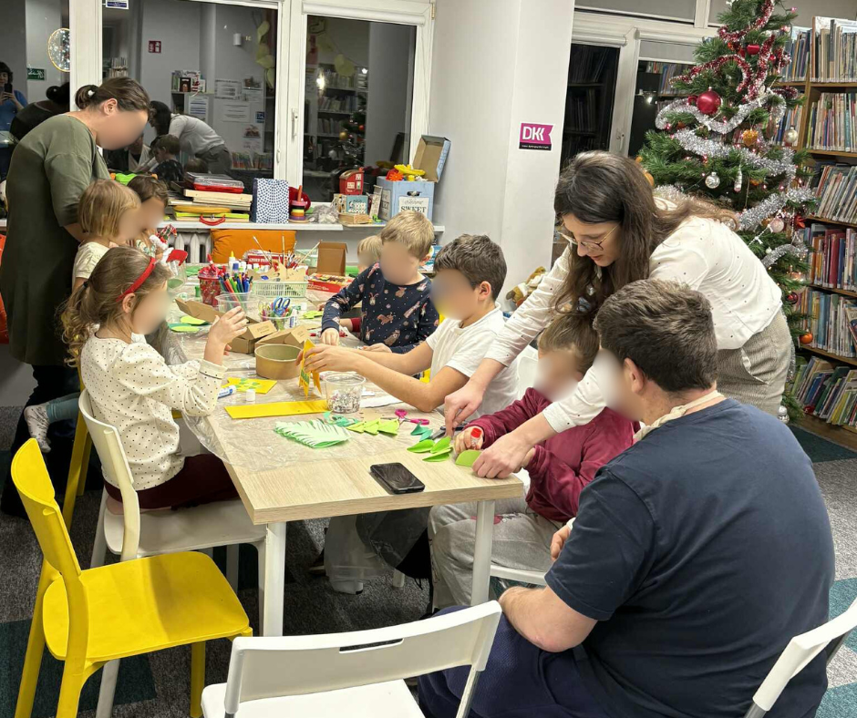 Szeroki kadr przedstawiający grupę dzieci i dorosłych wspólnie uczestniczących w warsztatach plastycznych. Wszyscy siedzą przy długim stole, kolorują, wycinają i kleją. W tle stoi udekorowana choinka świąteczna z bombkami i łańcuchami, co tworzy ciepłą, świąteczną atmosferę wspólnej zabawy i twórczości.