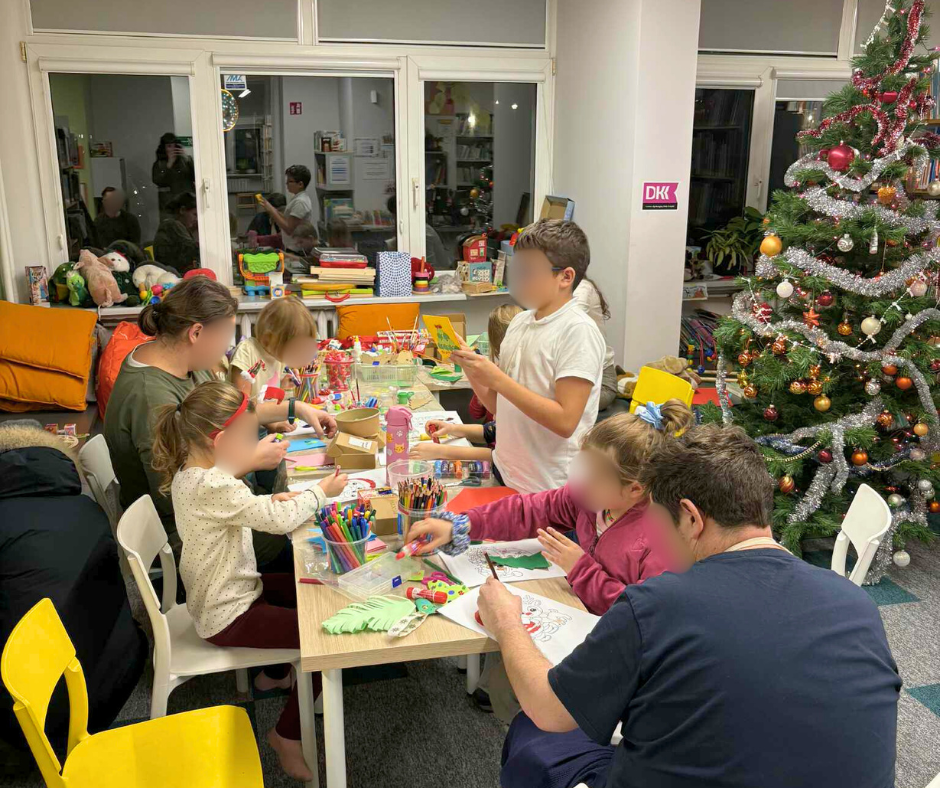 Szeroki kadr przedstawiający grupę dzieci i dorosłych wspólnie uczestniczących w warsztatach plastycznych. Wszyscy siedzą przy długim stole, kolorują, wycinają i kleją. W tle stoi udekorowana choinka świąteczna z bombkami i łańcuchami, co tworzy ciepłą, świąteczną atmosferę wspólnej zabawy i twórczości.