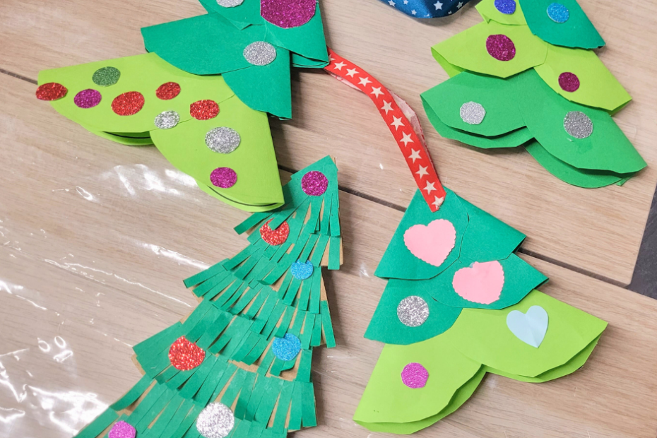 Kilka gotowych papierowych choinek leży na stole. Choinki są wykonane z różnych odcieni zielonego papieru i ozdobione kolorowymi, brokatowymi kółkami oraz papierowymi serduszkami. Każda praca jest inna, co podkreśla indywidualną kreatywność dzieci.