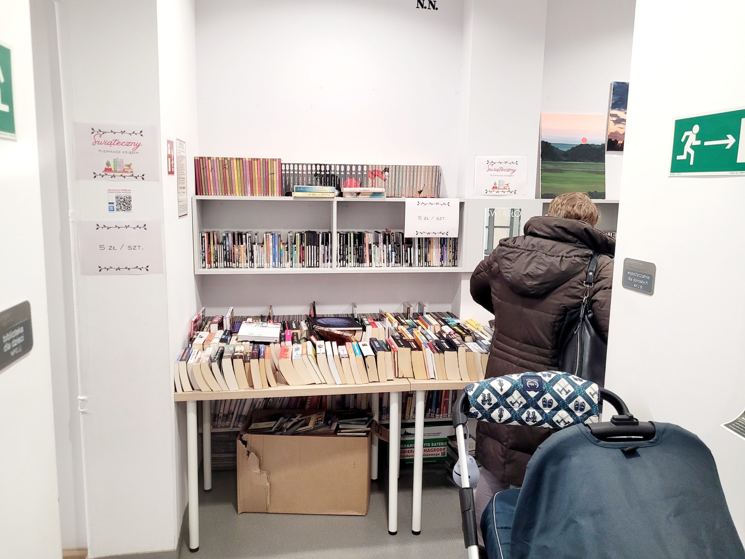Regały i stoły z używanymi książkami w bibliotece podczas świątecznego kiermaszu; kartki informują o cenie 5 zł za sztukę, obok widać wózek dziecięcy i osobę przeglądającą książki.