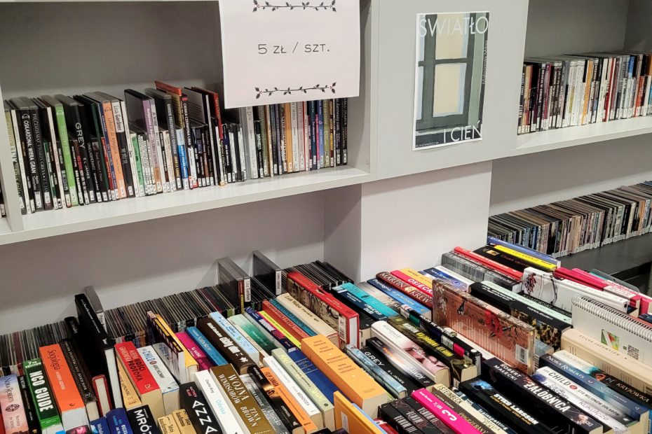 Zbliżenie na stoły gęsto zastawione książkami w różnych formatach; na regałach nad nimi plakaty „Świąteczny kiermasz książek” i informacja 5 zł / szt.