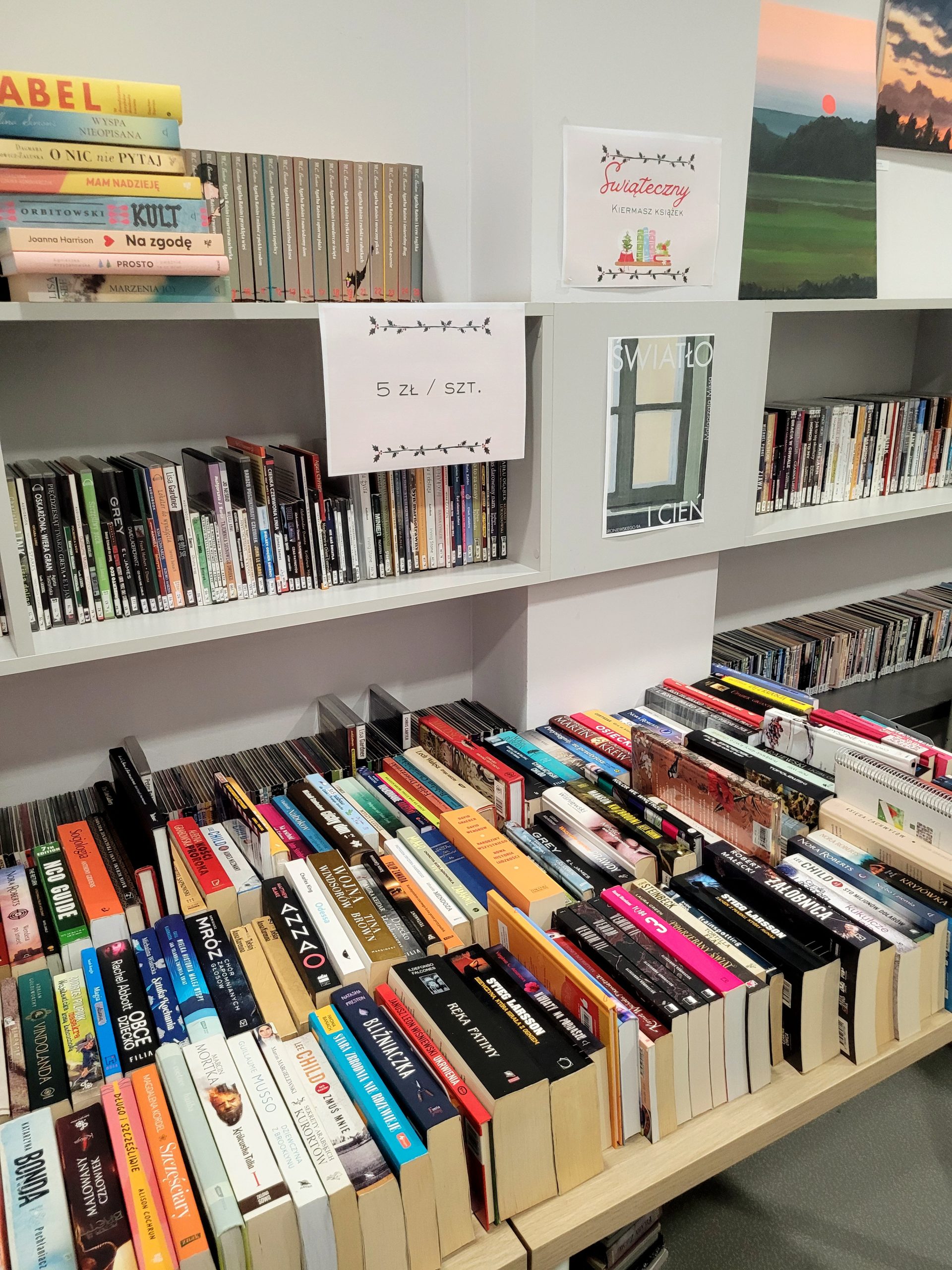 Zbliżenie na stoły gęsto zastawione książkami w różnych formatach; na regałach nad nimi plakaty „Świąteczny kiermasz książek” i informacja 5 zł / szt.
