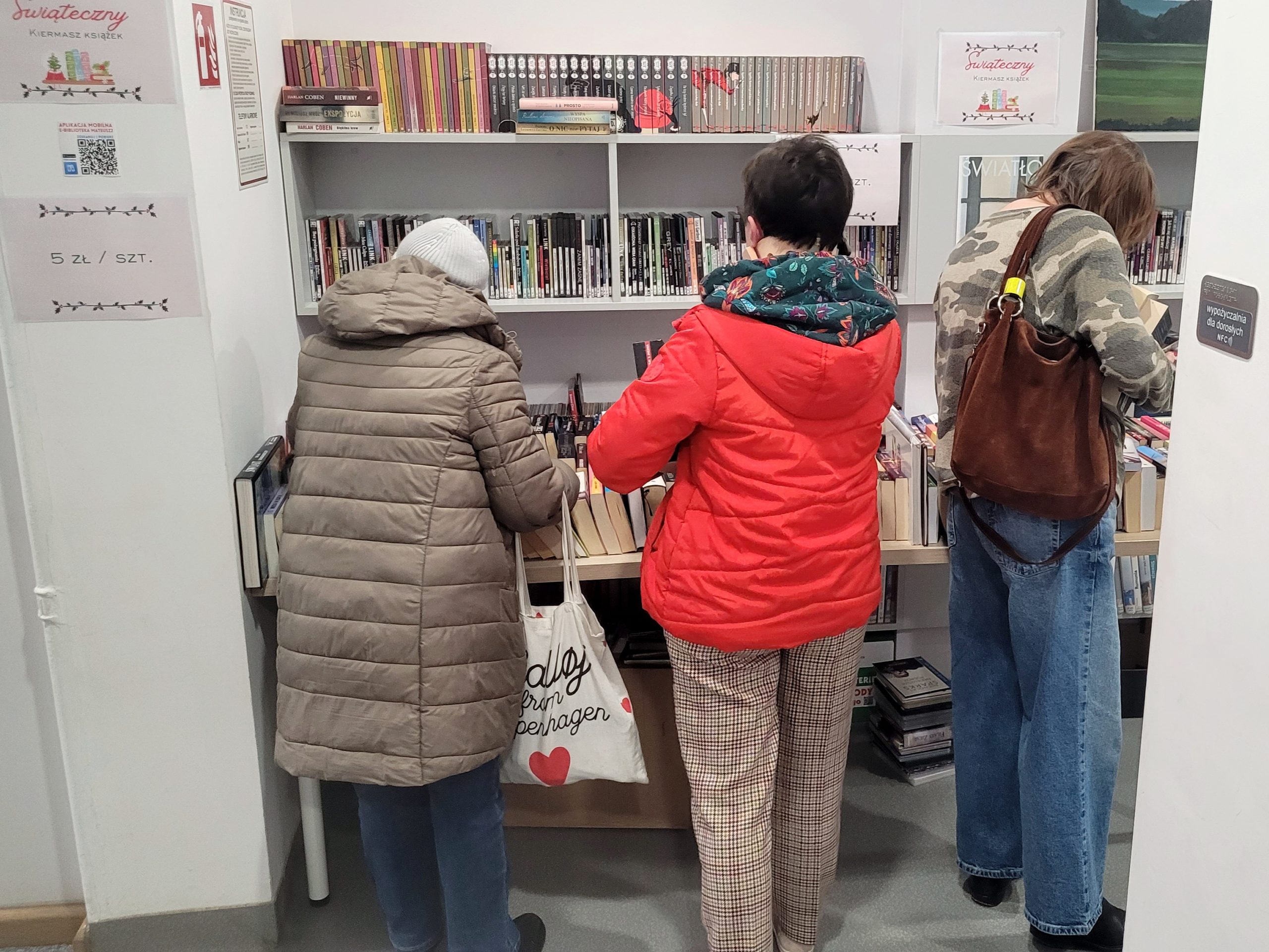 Trzy osoby stoją tyłem i przeglądają książki na stołach podczas kiermaszu w bibliotece; za nimi regały z książkami i oznaczenia cenowe.